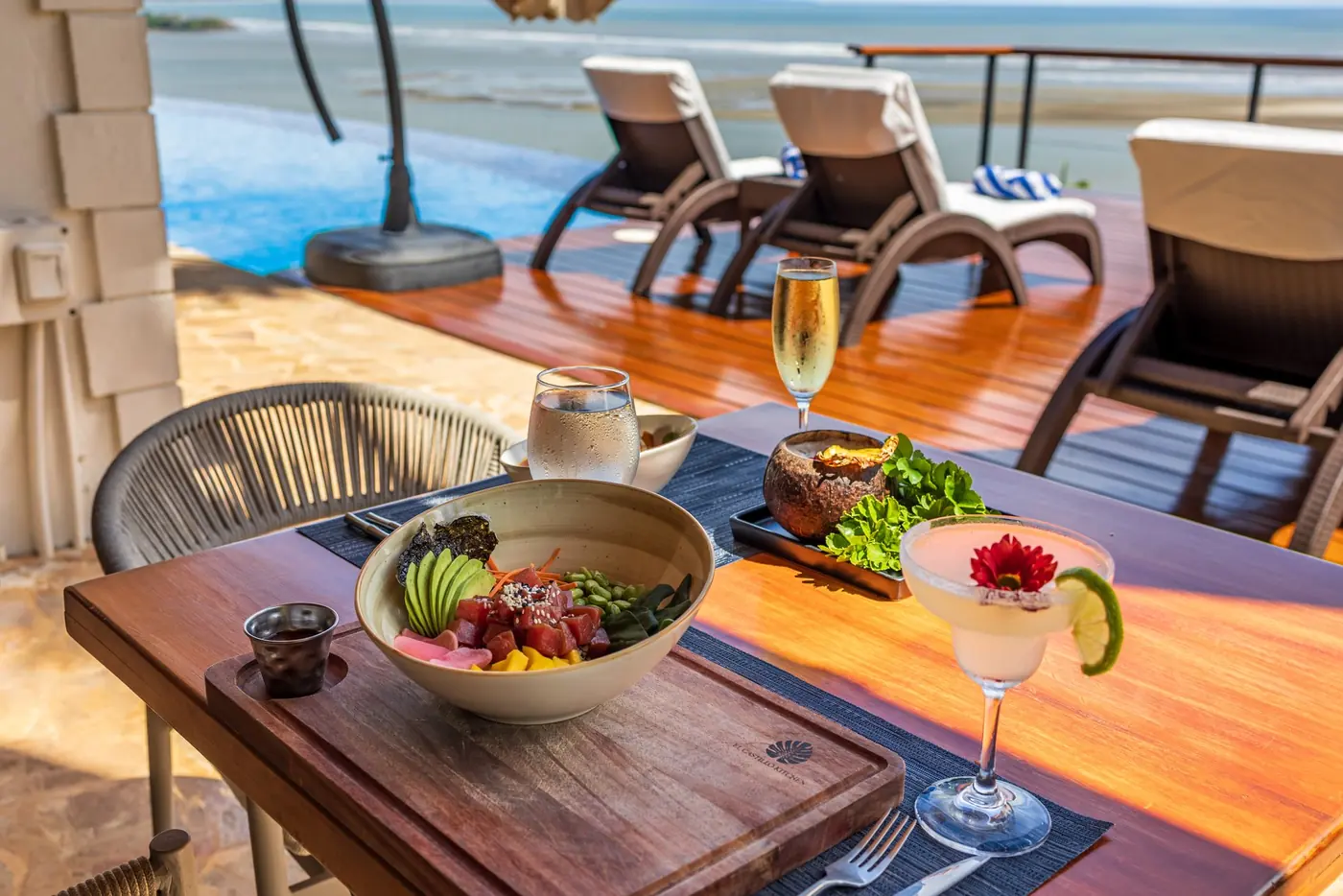 A poke bowl, cocktail and champagne on a sunlit table above the pool and ocean